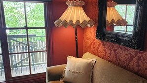 Una habitación con piso de madera, un sofá con cojines a rayas, una lámpara de pie alta, una ventana con vista a una terraza de madera y una pared con estampado.