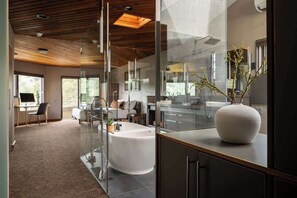 A modern hotel room with a glass partition, a white bathtub, a wooden ceiling, and a large vase on the counter.