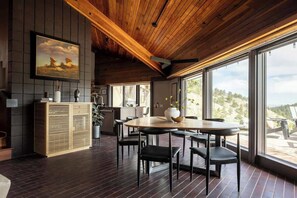Ein modernes Interieur mit Holzboden, einem Essbereich mit rundem Tisch und Stühlen, einer Küchenzeile und großen Fenstern mit Blick ins Freie.