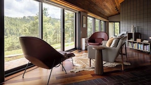 A modern interior with large windows, a wooden ceiling, a leather chair, a white sofa, and a bookshelf. - Architects Dream Home | Secluded Retreat | 30 Day (Golden)