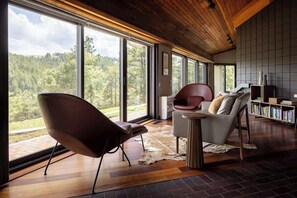 A modern interior with large windows, a wooden ceiling, a leather chair, a white sofa, and a bookshelf.