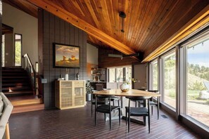 A modern kitchen with a wooden ceiling, a dining area with a round table and chairs, and large windows offering a view of the outdoors.