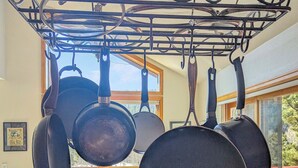 Een keuken met hangende potten en pannen, een eettafel met stoelen en uitzicht op het buitenleven door grote ramen.