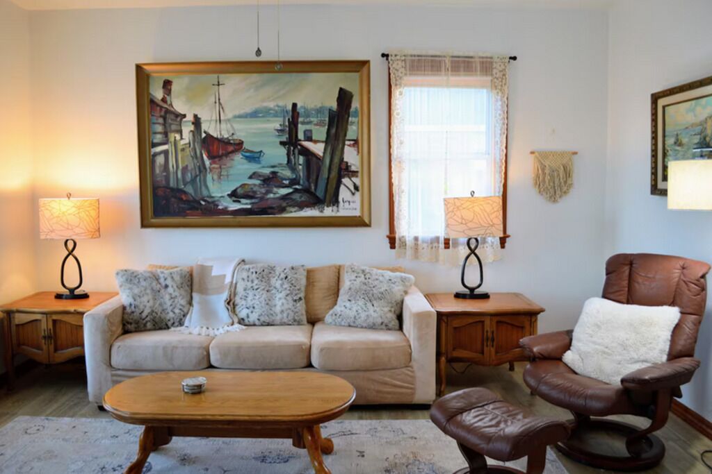 Un salon avec un canapé beige, une table basse en bois, un fauteuil en cuir marron et deux lampes. Un grand tableau représentant un paysage portuaire est accroché au mur.
