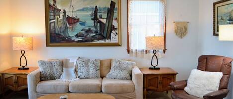Un salon avec un canapé beige, une table basse en bois, un fauteuil en cuir marron et deux lampes. Un grand tableau représentant un paysage portuaire est accroché au mur.