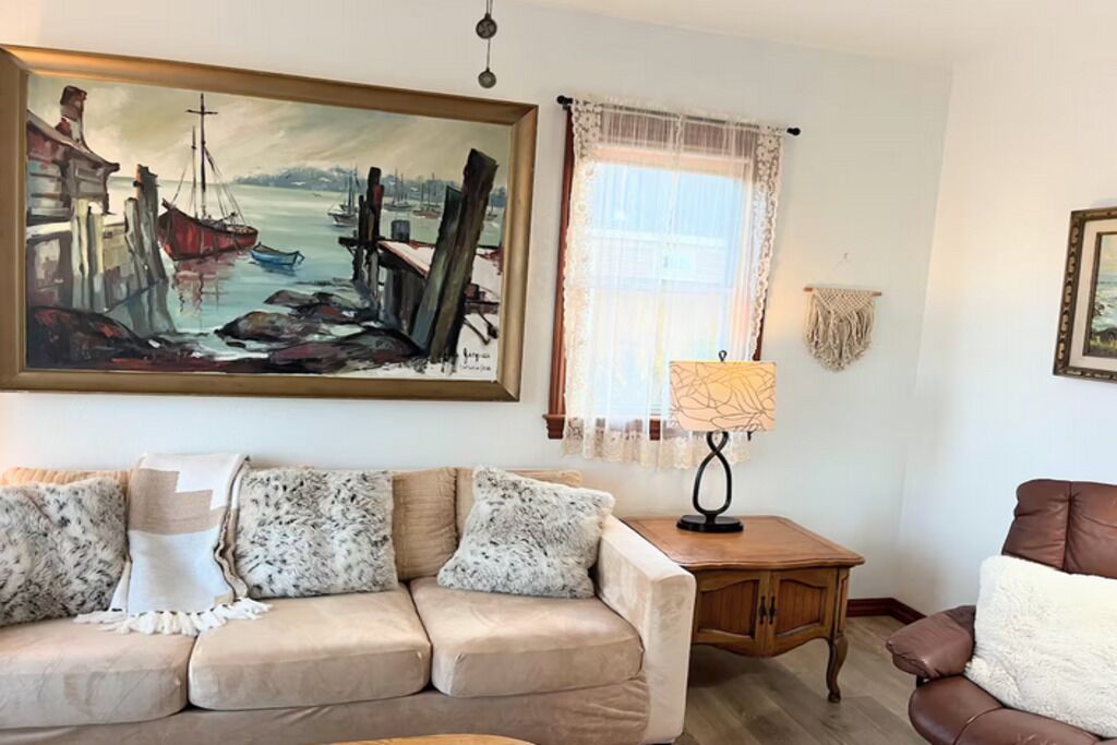 Uma sala de estar com um sofá bege, uma mesa lateral de madeira, uma luminária, um quadro emoldurado com uma paisagem portuária e uma janela com cortinas leves.