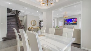 A modern dining area with a glass table and white chairs, a staircase, and a television in the background.