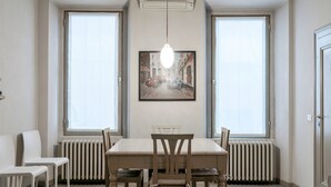 Un coin repas comprenant une table, des chaises et un luminaire suspendu. Il y a deux fenêtres équipées de stores, un radiateur et un tableau encadré accroché au mur.