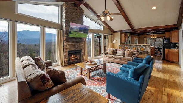 A spacious living room with a fireplace, large windows, and a kitchen area. - Views, Hot tub, Pool Table! (Sevierville)