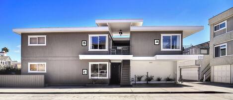 A modern two-story residential building with a flat roof, a covered front porch, and a small front yard with plants.