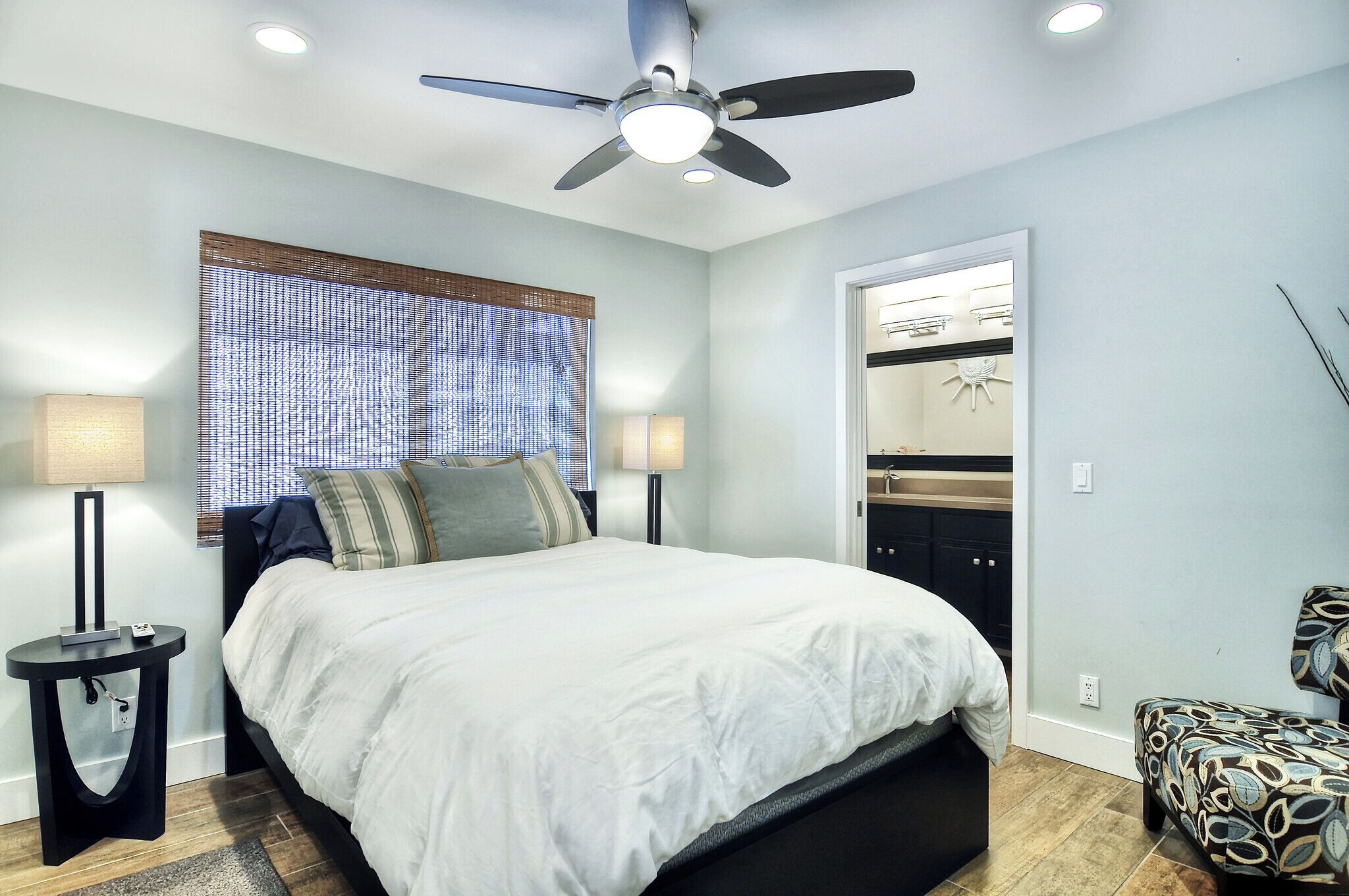Une chambre à coucher avec un lit, deux tables de chevet, un ventilateur de plafond, une fenêtre avec des stores, et une porte de salle de bain visible au fond.