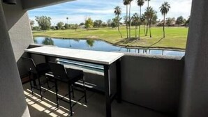 A balcony with a table and chairs, overlooking a golf course with palm trees and a pond.