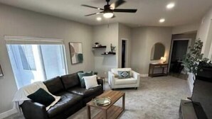 A spacious living room with a black leather sofa, a white armchair, and a wooden coffee table. There is a ceiling fan, a window with blinds, and a door leading to another room.