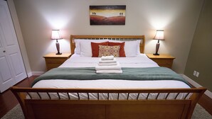 Un lit bien fait, avec du linge de lit blanc, un plaid vert et une pile de serviettes blanches. Deux lampes de chevet sont posées sur des tables de nuit en bois, et un tableau est accroché au-dessus du lit.