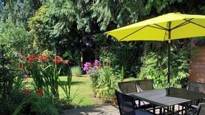 Une terrasse extérieure avec une table et des chaises, entourée d’une végétation luxuriante et de plantes en fleurs.