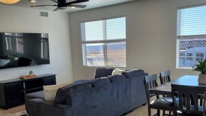 Ein modernes Wohnzimmer mit großem Flachbildfernseher, einem dunkelblauen Ecksofa, einem Esstisch mit Stühlen und einem Deckenventilator.