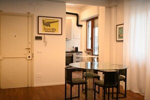 Une pièce avec une table en verre et quatre chaises, une porte blanche et, au fond, un coin cuisine.
