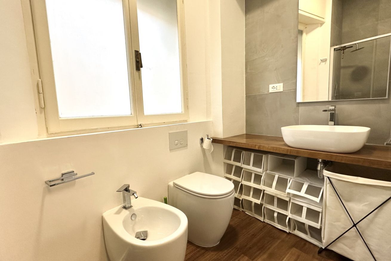 A bathroom with a toilet, bidet, sink, mirror, and storage baskets.