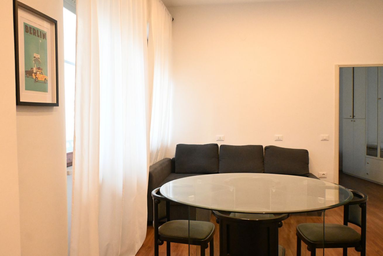 Een kamer met een ronde glazen tafel, vier stoelen, een bank met kussens, een ingelijst schilderij aan de muur, en een deur op de achtergrond.
