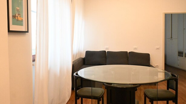 Une pièce avec une table ronde en verre, quatre chaises, un canapé avec des coussins, un tableau encadré au mur et une porte au fond.