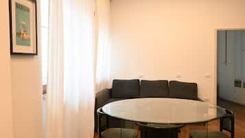 Een kamer met een ronde glazen tafel, vier stoelen, een bank met kussens, een ingelijst schilderij aan de muur, en een deur op de achtergrond.