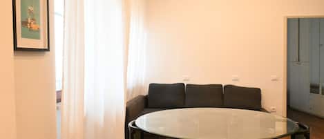 Una habitación con una mesa redonda de vidrio, cuatro sillas, un sofá con cojines, un cuadro en la pared y una puerta al fondo.