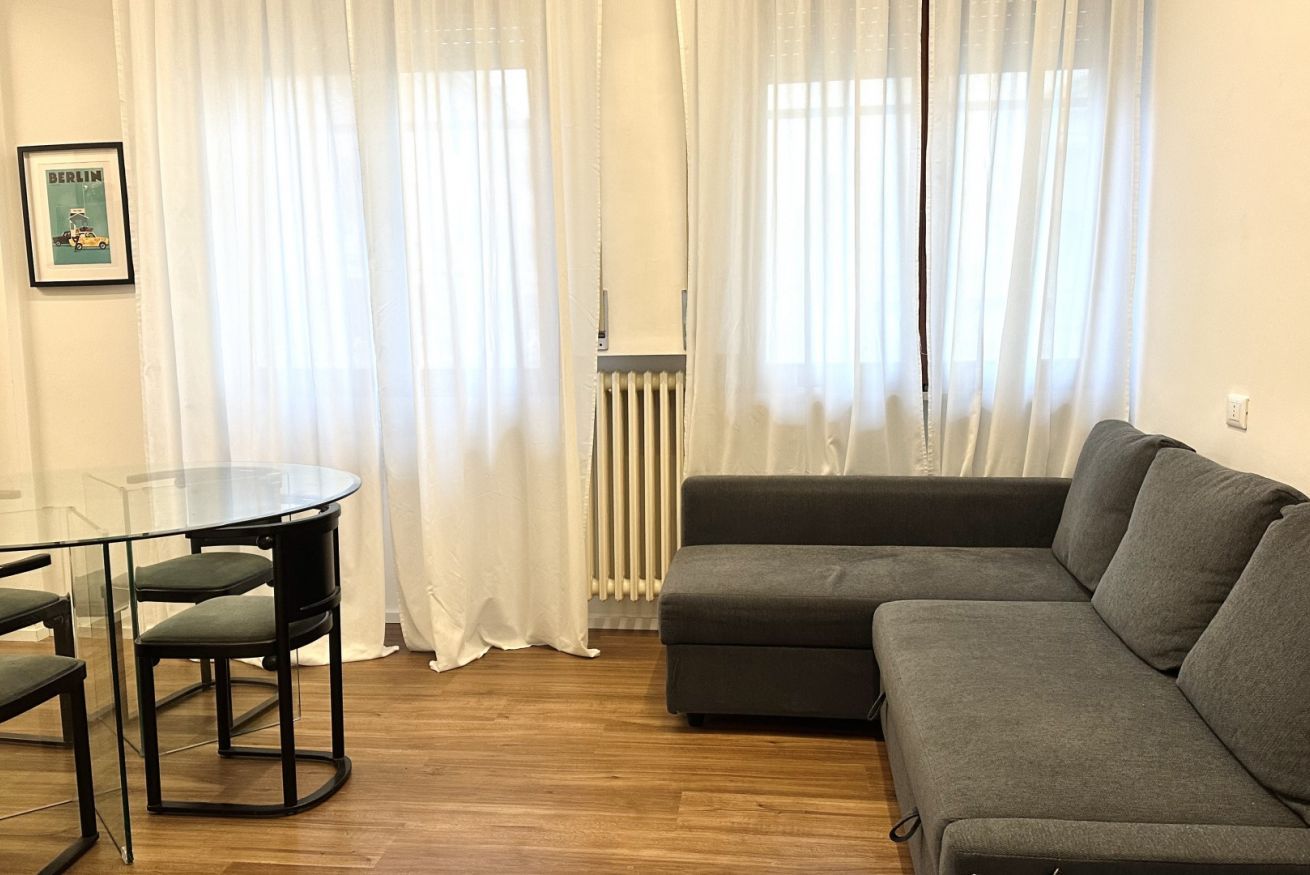 A living room with a grey sofa, a glass table with chairs, and white curtains.