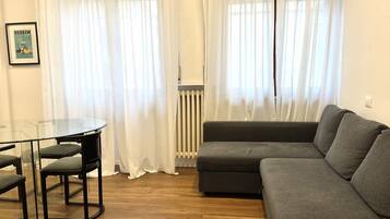 A living room with a grey sofa, a glass table with chairs, and white curtains.