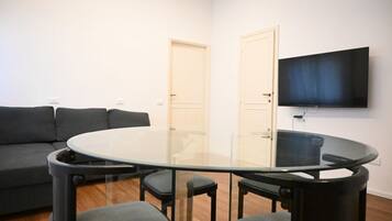 A living room with a glass-top table, four chairs, a sofa, and a flat-screen TV mounted on the wall.