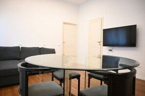 Un salon comprenant une table avec un plateau en verre, quatre chaises, un canapé et un téléviseur à écran plat fixé au mur.