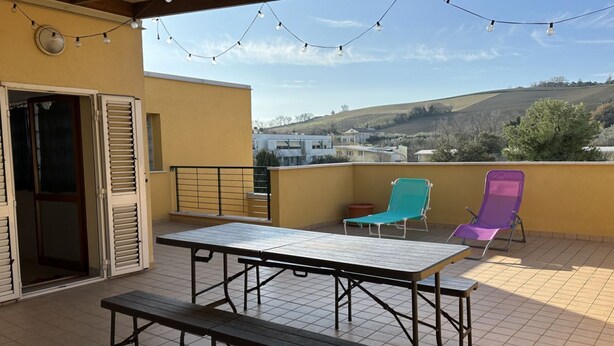Una terrazza con tavolo e panche, una porta con persiane e vista su un paesaggio collinare.