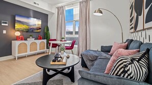 A modern living room with a sofa, a coffee table, and a TV mounted on the wall. There is a dining table with chairs and a window with curtains.