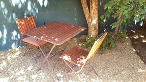 Una mesa de madera y dos sillas en un espacio al aire libre sombreado, con un árbol y una pared al fondo.