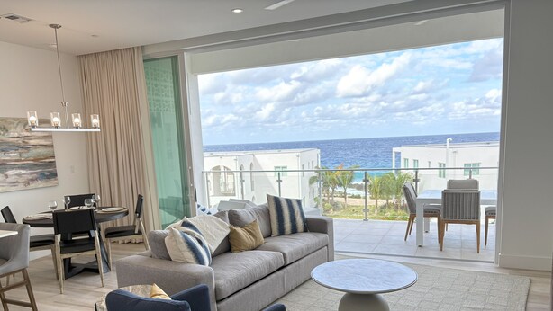 A modern living room with a grey sofa, blue armchairs, and a round coffee table. A dining area with a table and chairs is visible, and a balcony with a view of the sea and buildings is also seen.
