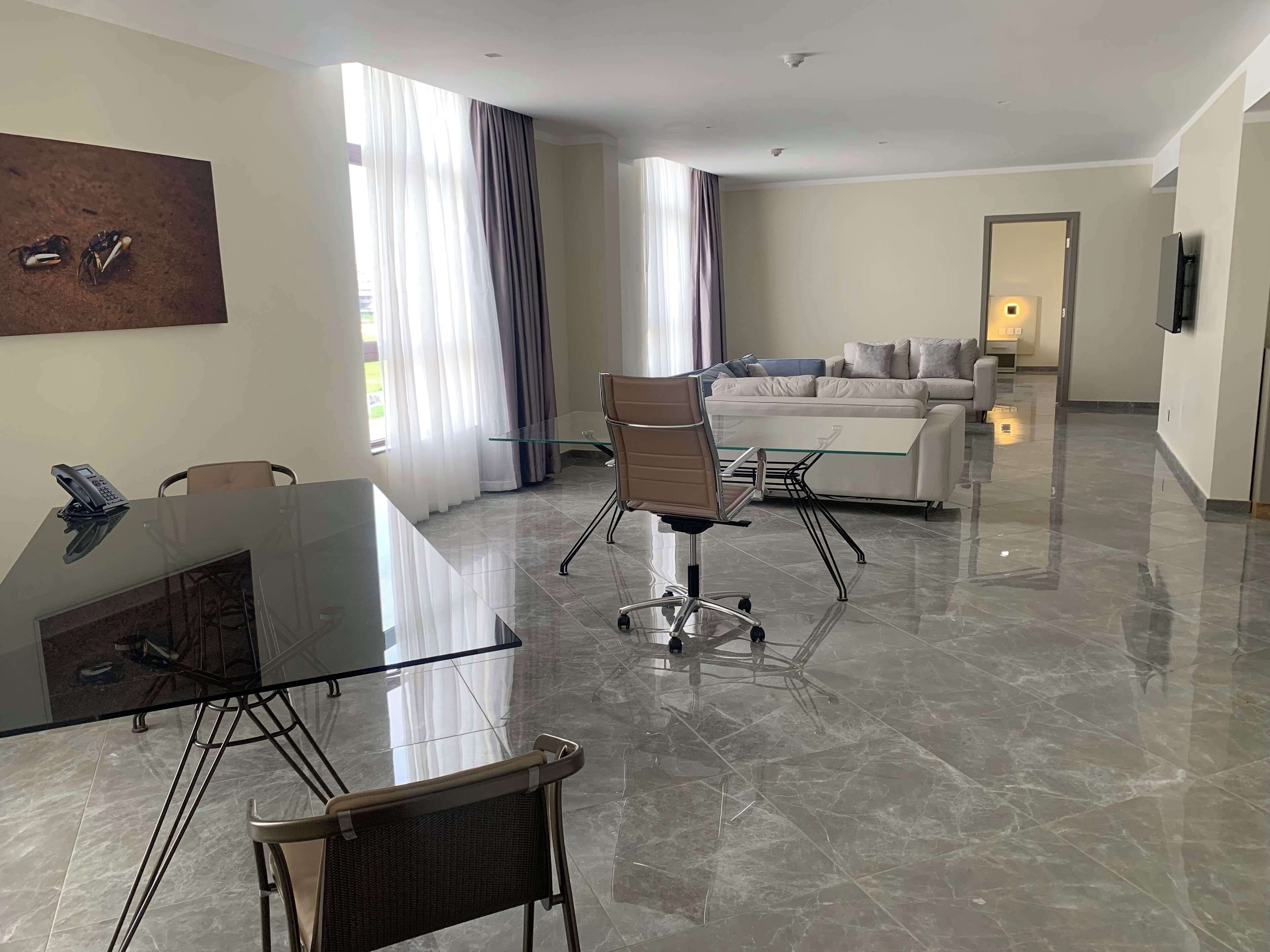 Een moderne hotelkamer met een glazen eettafel, een bank en een groot raam met gordijnen.