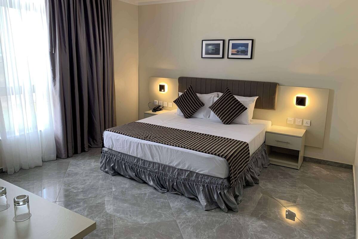 A modern hotel room with a large bed, two bedside tables, a nightstand, and a small desk.