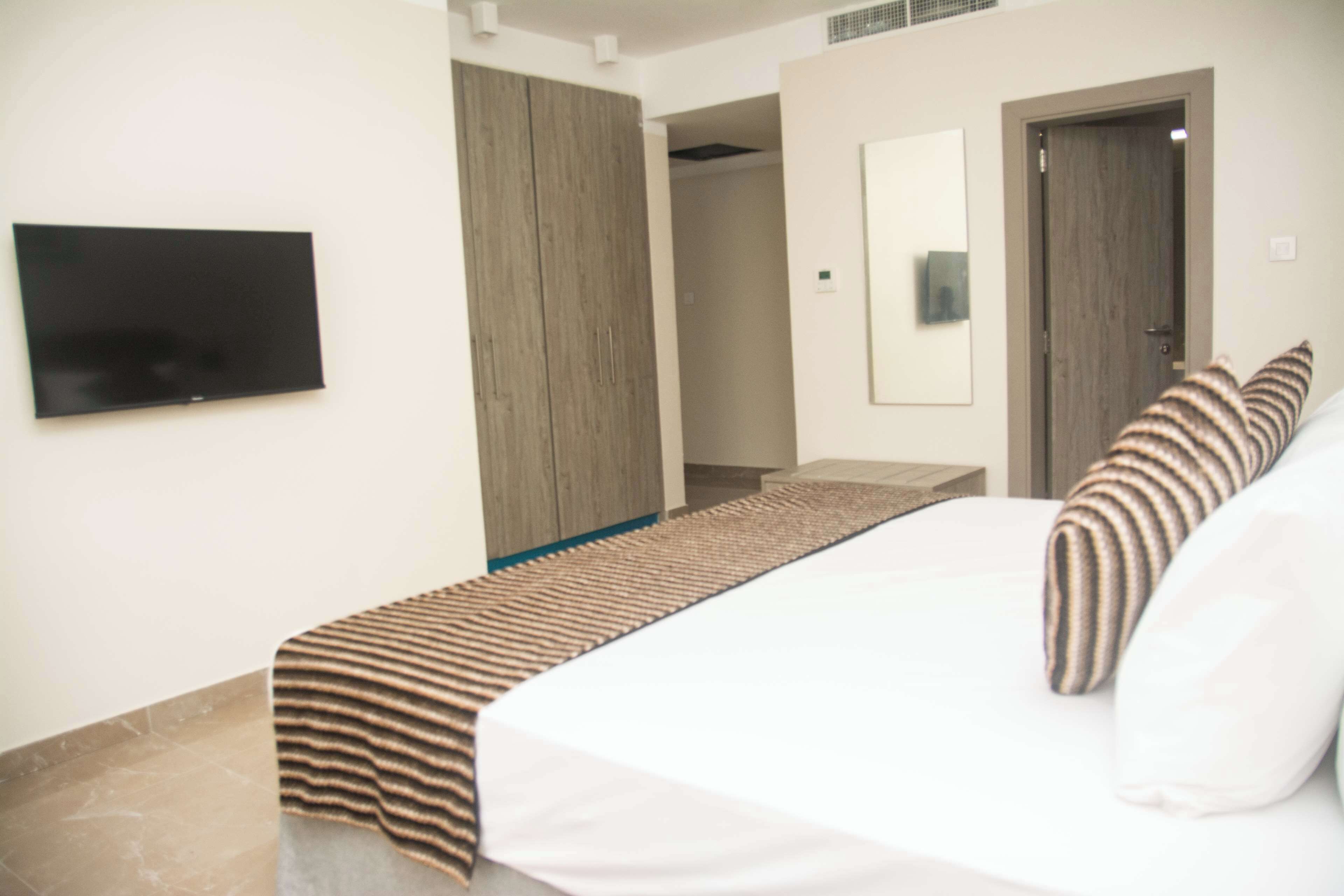 A hotel room with a bed, a television mounted on the wall, a wooden wardrobe, and a door.