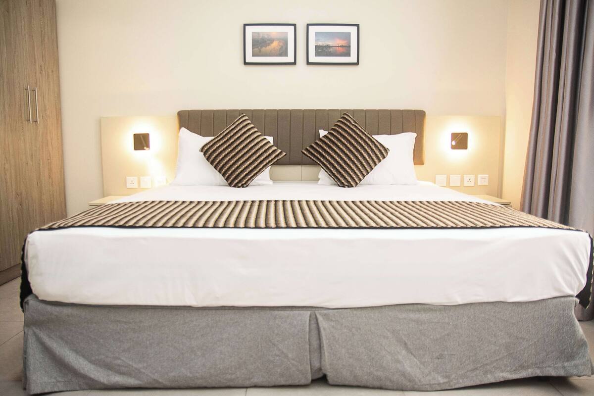 A hotel room with a large bed, two bedside tables, a headboard with integrated lighting, and two framed pictures on the wall.