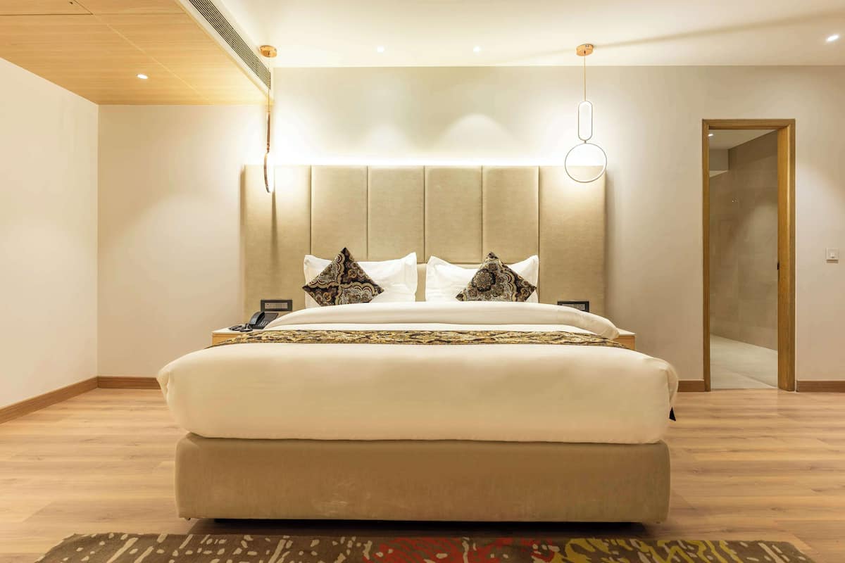 Quarto de hotel moderno com uma cama grande, cabeceira com iluminação integrada, mesa de cabeceira com telefone e piso de madeira.
