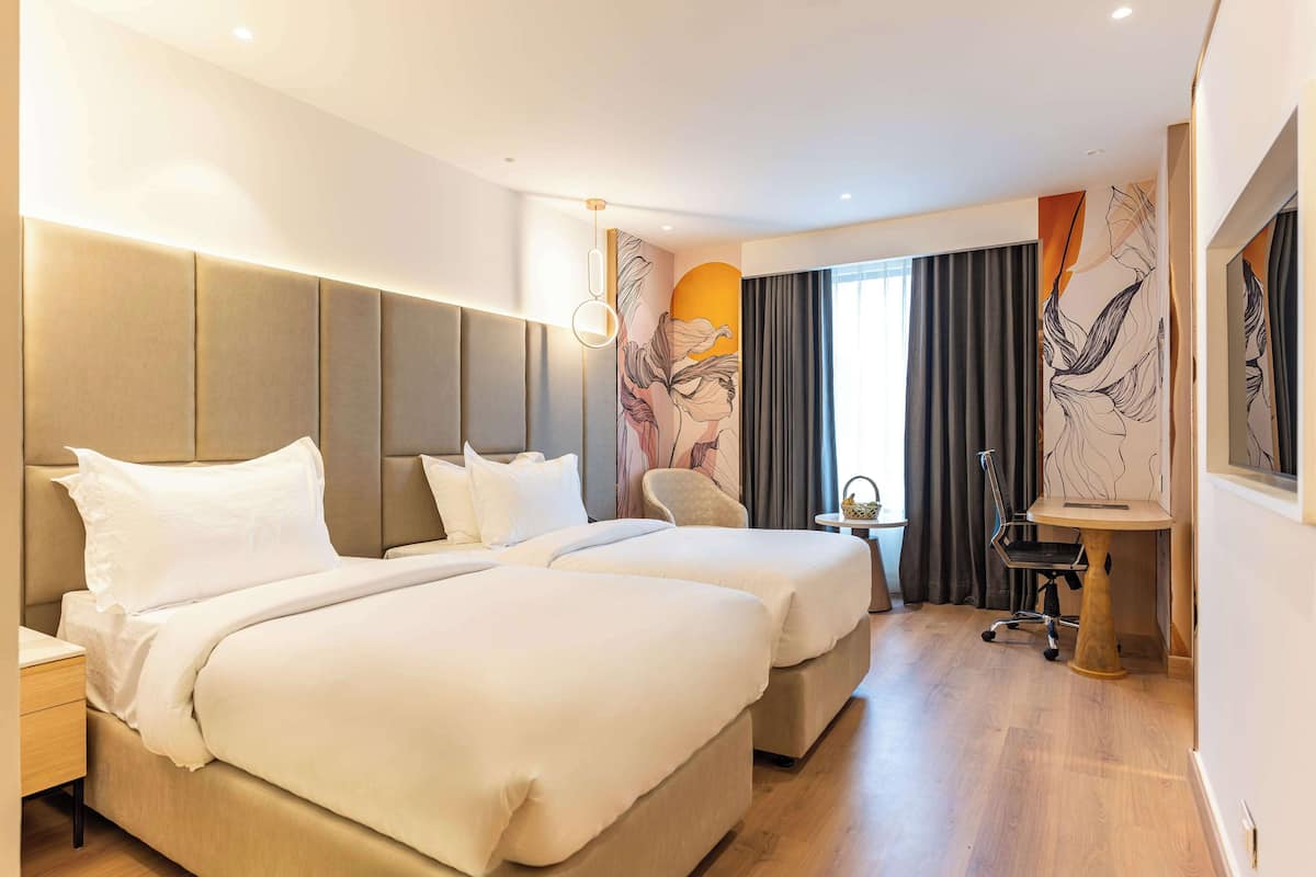 Quarto de hotel com duas camas, uma escrivaninha, uma cadeira e uma janela grande com cortinas.