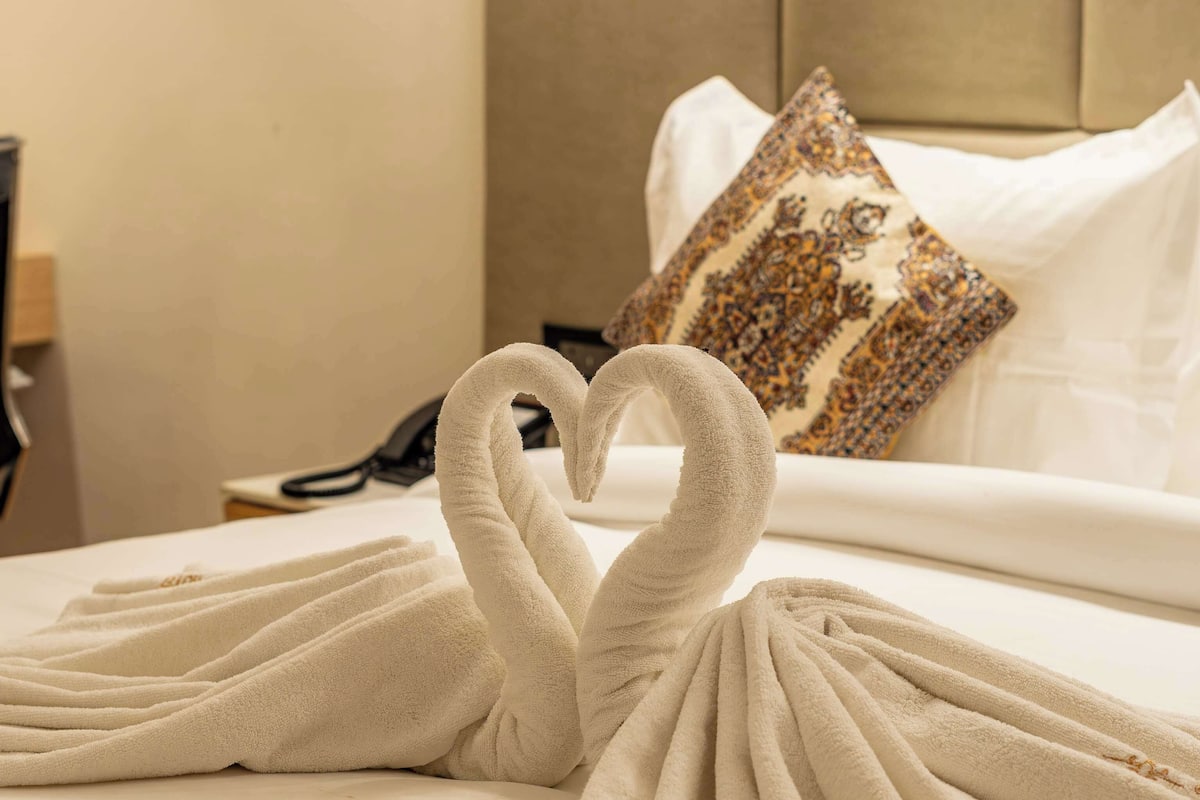 Quarto de hotel com cama bem arrumada, arranjo de toalhas em formato de coração e abajur.