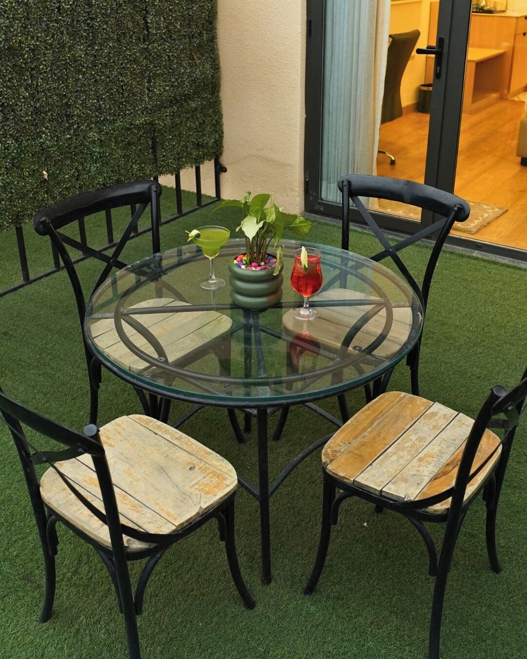 A round glass table with four wooden chairs, a potted plant, and two drinks.