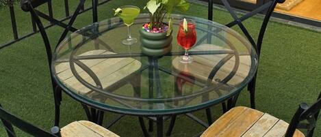 A round glass table with four wooden chairs, a potted plant, and two drinks.