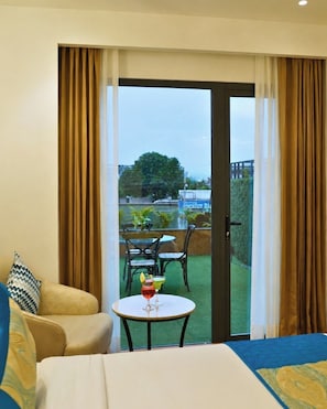 Une chambre d’hôtel avec un lit, une chaise, une petite table avec des boissons et une vue sur un espace extérieur verdoyant et un bâtiment.