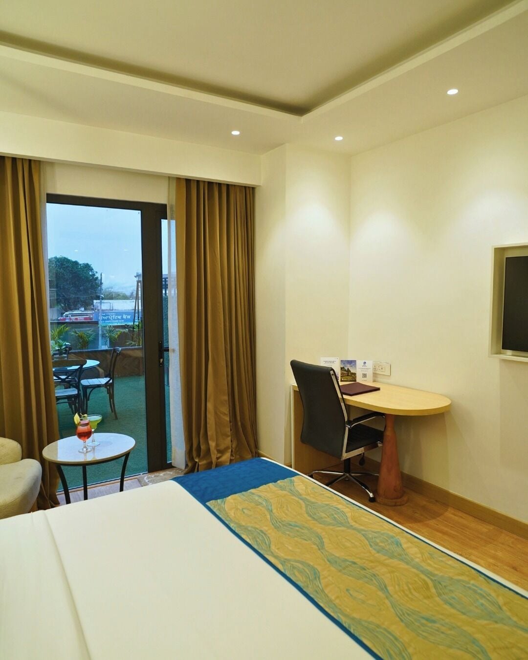 A hotel room with a bed, a desk, a chair, a small table with a drink, and a view of the outside.