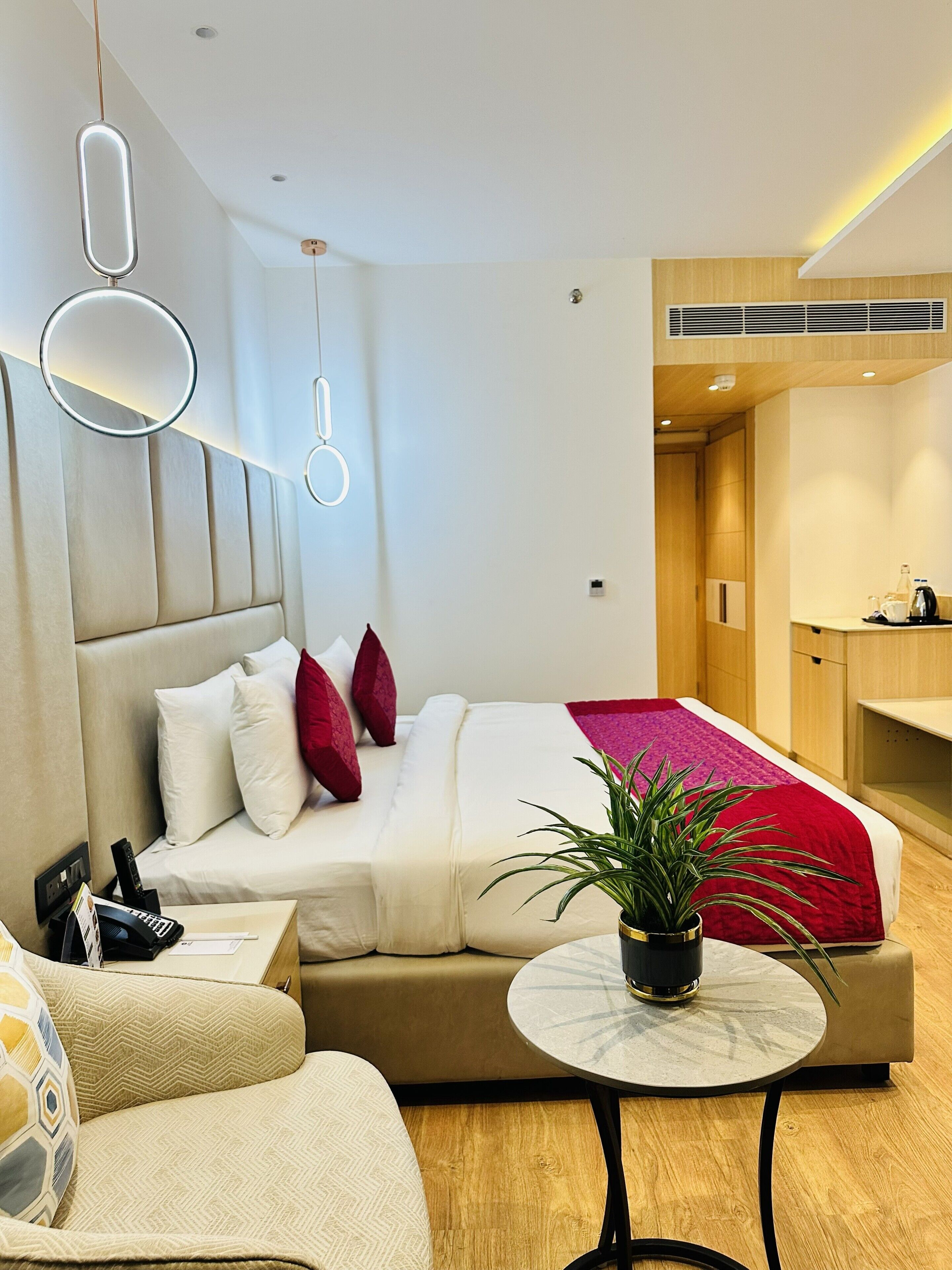 Una habitación de hotel moderna con una cama grande, una mesita de noche con teléfono y lámpara, una mesa redonda con una planta, y una zona de kitchenette.