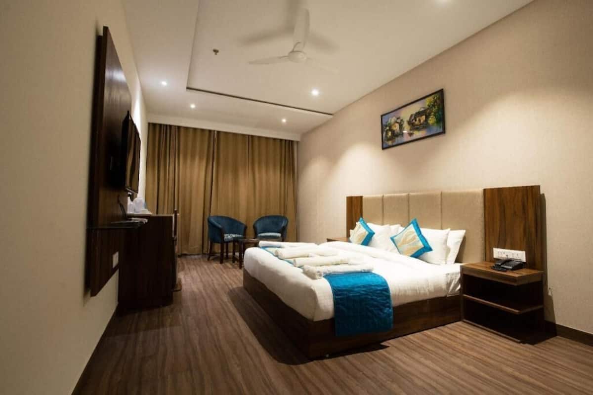 Quarto de hotel com cama, escrivaninha, duas cadeiras, televisão, ventoinha no teto e um quadro na parede.