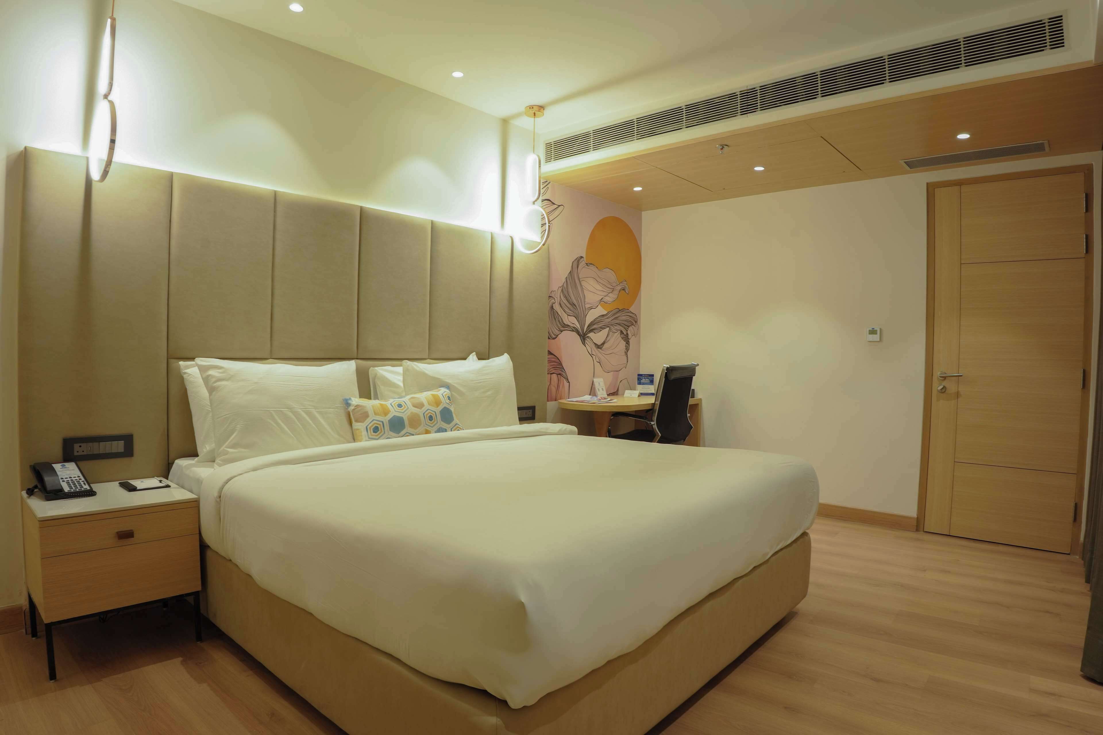 A hotel room with a large bed, bedside table with a phone, a desk with a chair, and a door.