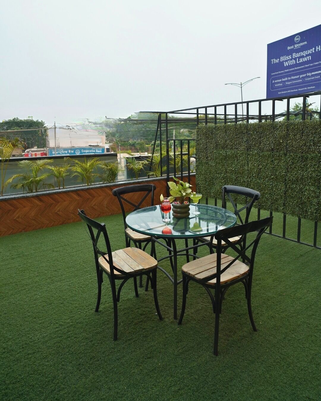 A round glass table with four chairs, a potted plant, and a cocktail on a rooftop terrace with artificial grass and a view of a cityscape.
