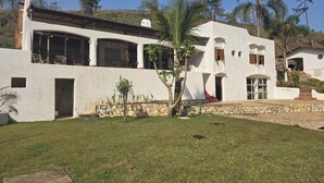 Una casa bianca a due piani con fondamenta in pietra, ampie finestre e un piccolo giardino anteriore con una palma.